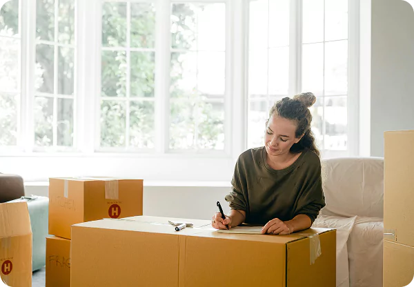 A woman sits at a table surrounded by boxes, writing with a pen.
