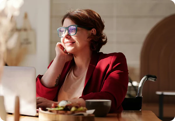 A woman with glasses is seated at a table, engaged with her laptop